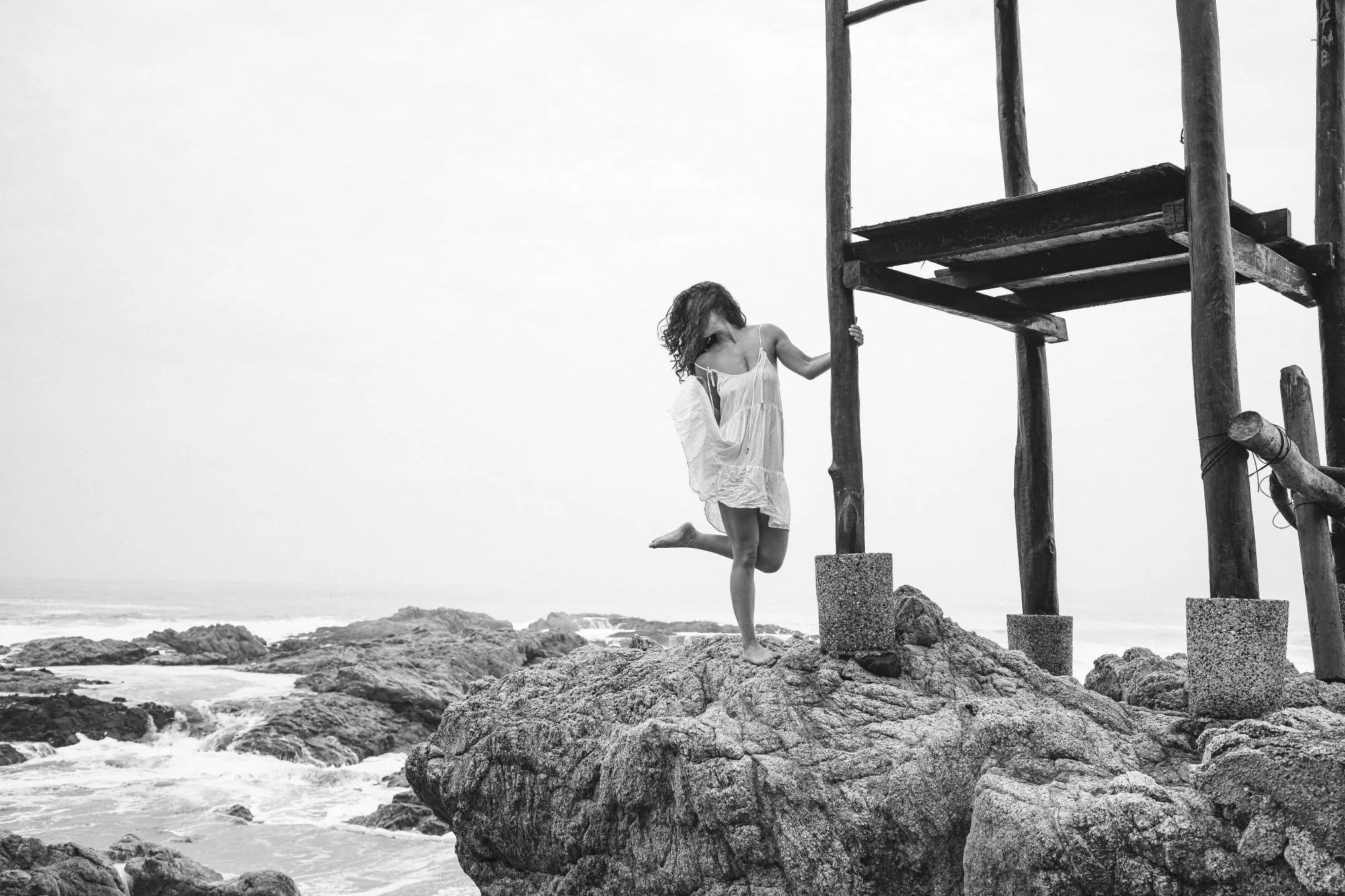 Retrato en blanco y negro con vestido blanco sobre las rocas junto a una torre de salvavidas de madera